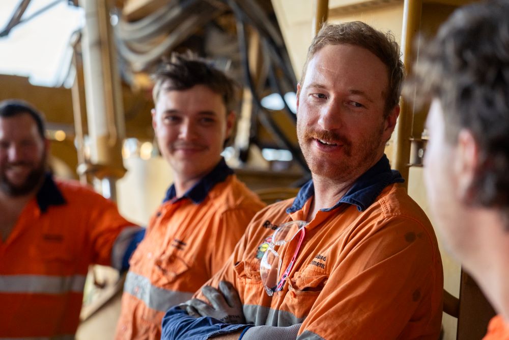 3 construction workers in orange and blue hig vis clothing standing up against machinery